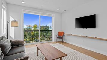 A modern living room with a sofa, a wooden coffee table, a TV mounted on the wall, and a balcony with a view of buildings and trees.