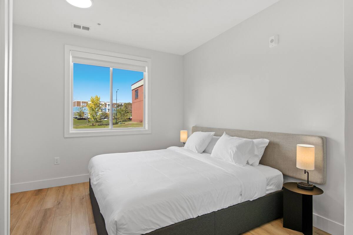 A modern bedroom with a large bed, a nightstand with a lamp, and a window offering a view of buildings and greenery.