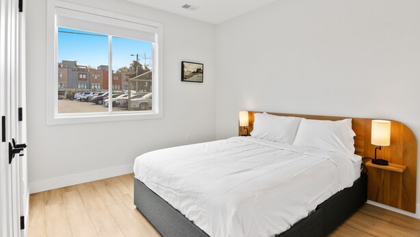 A neatly made bed with white linens in a room with a large window, a framed picture on the wall, and a nightstand with a lamp. - The Loop Downtown By Kasa (Traverse City)
