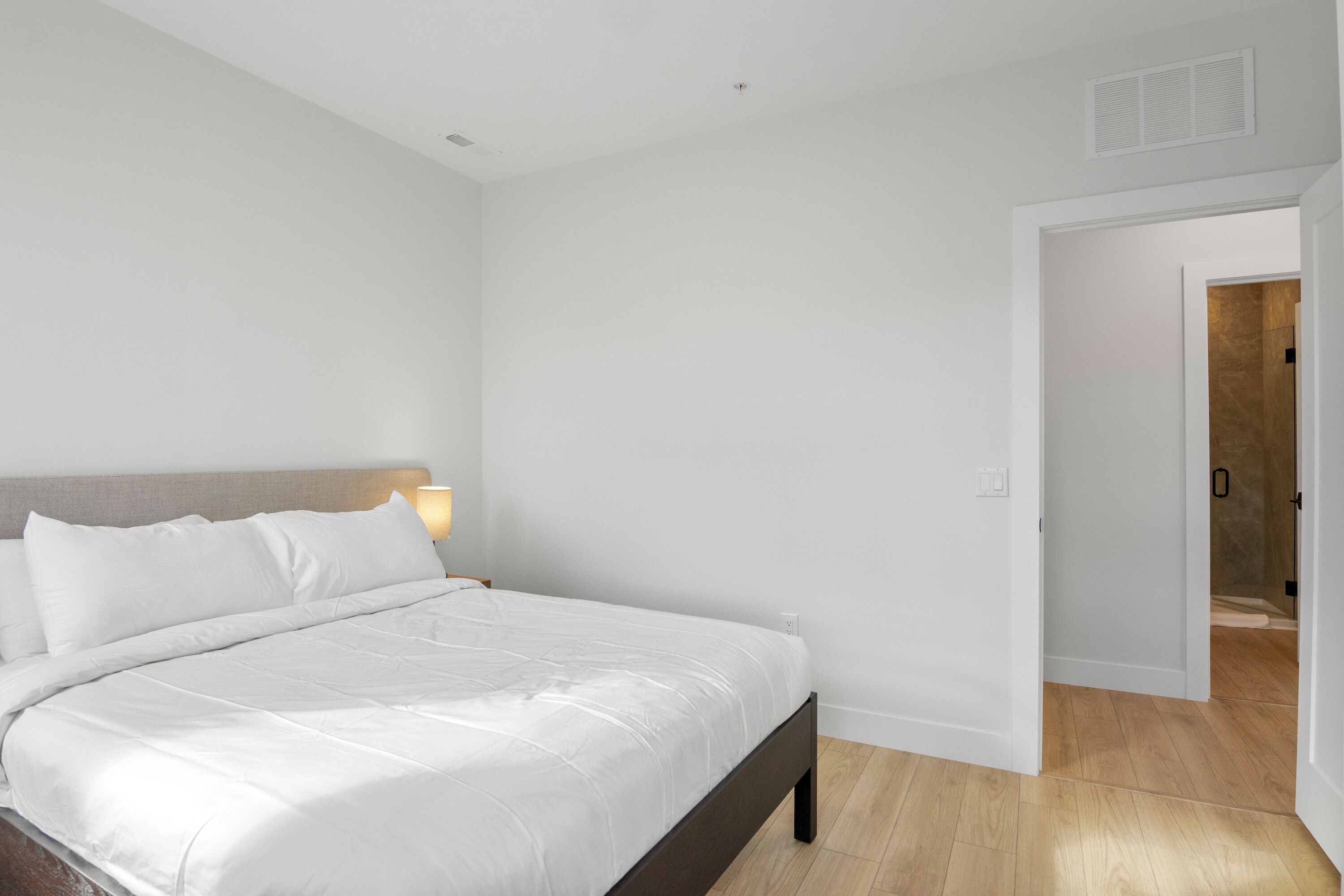 A bedroom with a bed, a nightstand with a lamp, and a door leading to a bathroom.