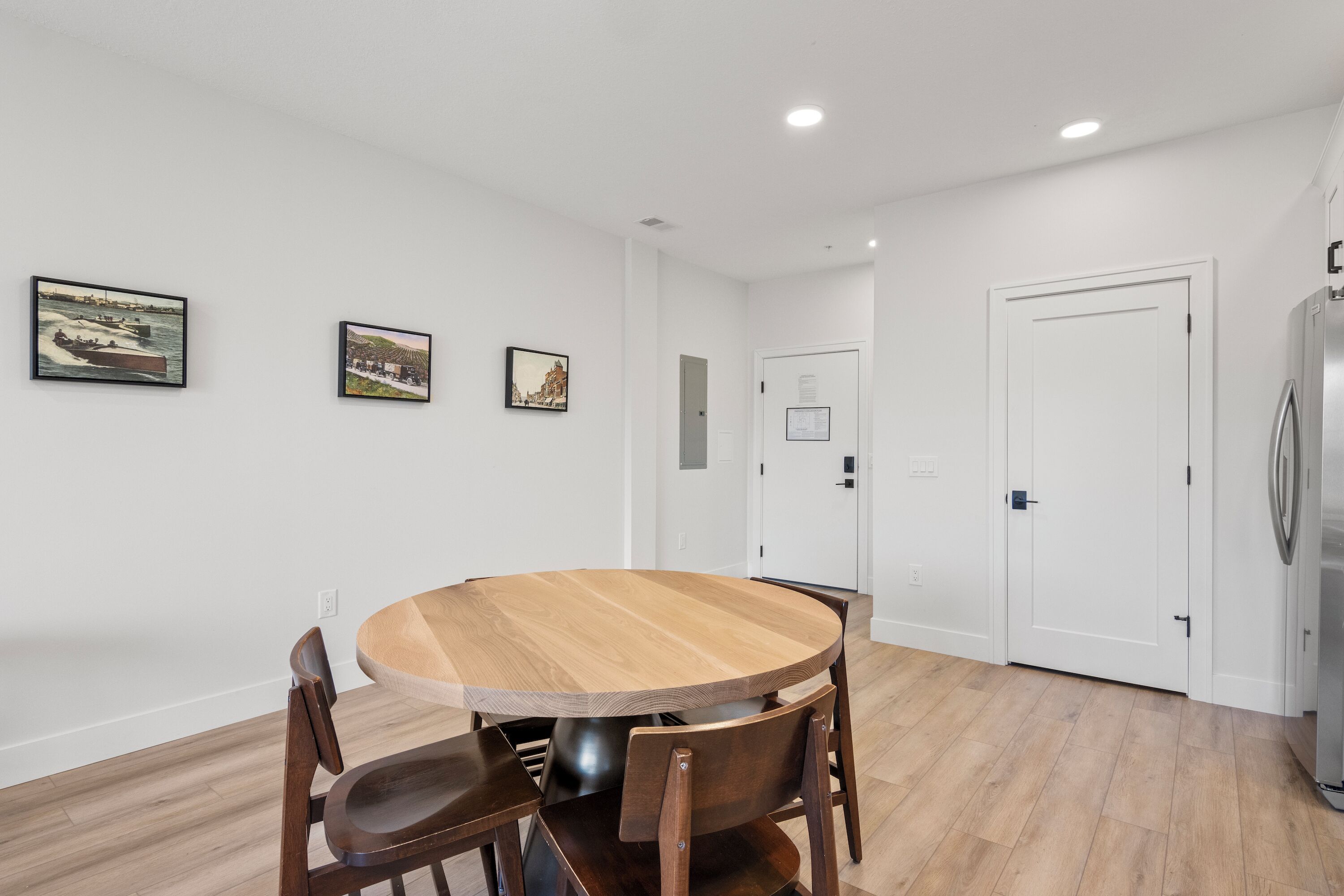 A dining area with a round wooden table and four chairs, white walls, and three framed pictures on the wall.