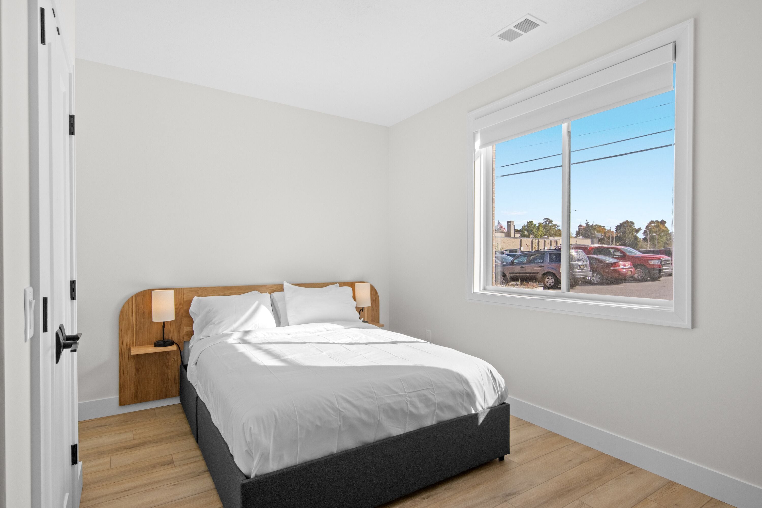 A bedroom with a bed, a nightstand, a window with a view of parked cars, and a door.
