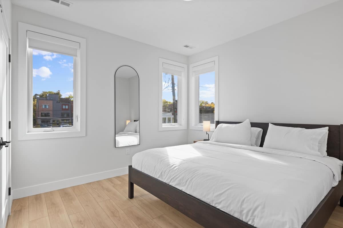 A bedroom with a large bed, two windows, a mirror, and a nightstand with a lamp.