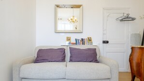 Un sofá de tonos neutros con dos cojines, un candelabro, una puerta con perchero y una pequeña estantería con libros.