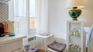 Una habitación pequeña con una mesa, dos sillas, una lámpara, un radiador, un estante con teteras y una ventana con cortinas translúcidas.