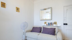 Un salón con un sofá gris, dos cojines morados, un ventilador de pie, una puerta blanca y una estantería con libros y fotos enmarcadas.