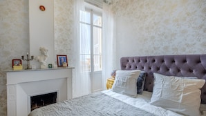 A bedroom with a fireplace, a bed with a tufted headboard, and a window with curtains. - Apartment 'Jolie Proche Monaco' with Wi-Fi (Beausoleil)