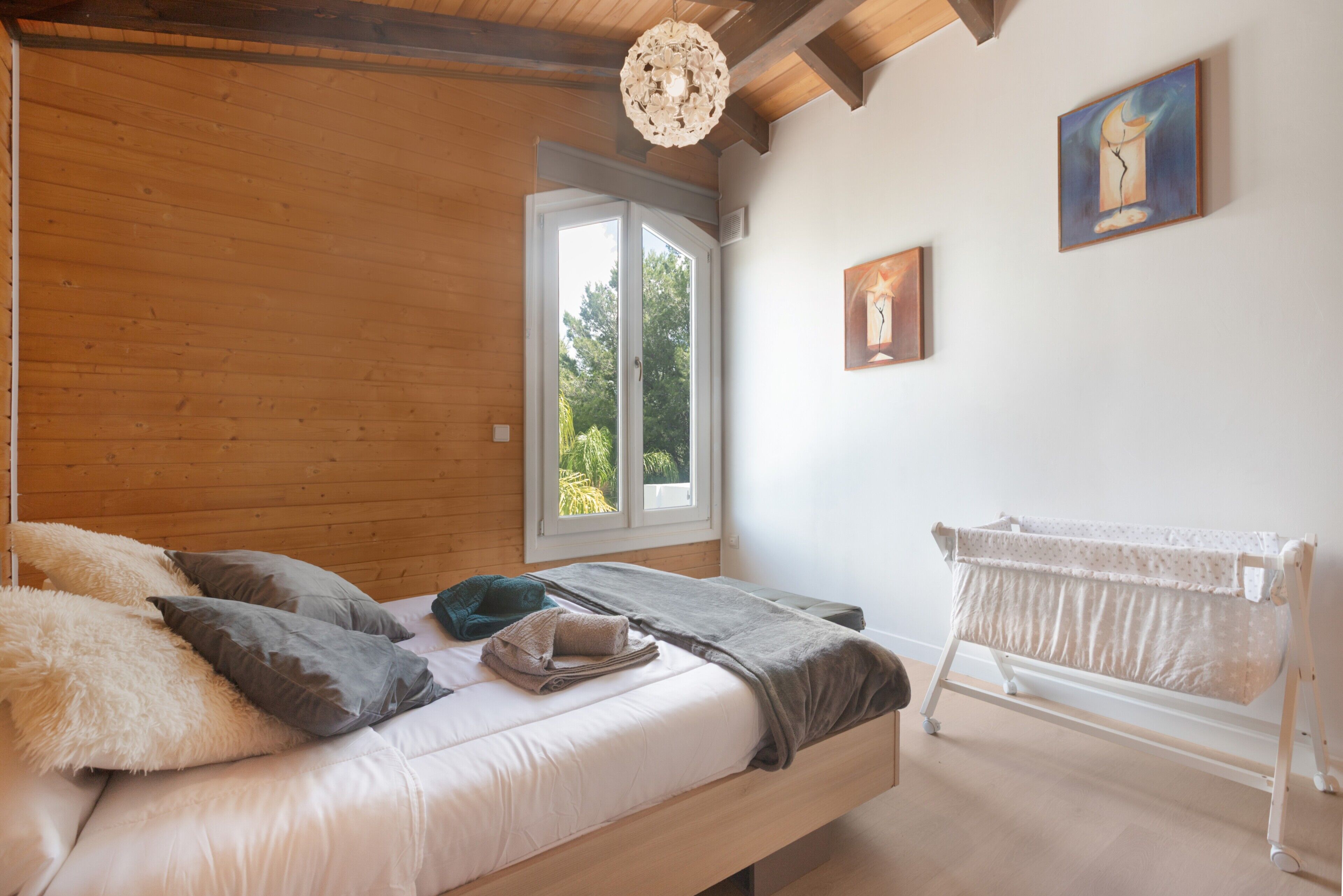 Un dormitorio con techo de madera, una cama con almohadas, una mesita de noche, una lámpara colgante, una ventana y una cuna.