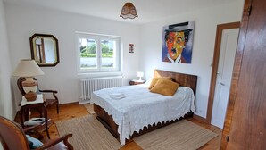 Una camera da letto con pavimento in legno, un letto con copripiumino bianco, una sedia, un tavolo con una lampada, una finestra e un quadro appeso al muro.