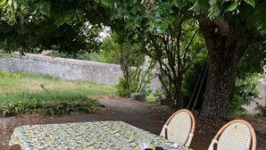 Un tavolo da pranzo all'aperto con tovaglia fantasia, circondato da sedie in vimini, in un ambiente naturale con alberi e un muro di pietra sullo sfondo.