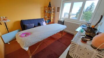 Een kamer met een massagetafel, een raam met uitzicht, een boekenkast en een klein tafeltje met schalen.