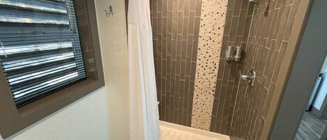 Une douche à l’italienne avec une porte en verre, des murs carrelés et une fenêtre avec des stores.