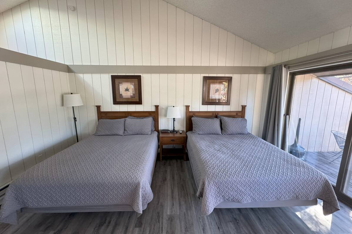A room with two beds, wooden headboards, a nightstand with a lamp, and a window with a view of the outdoors.