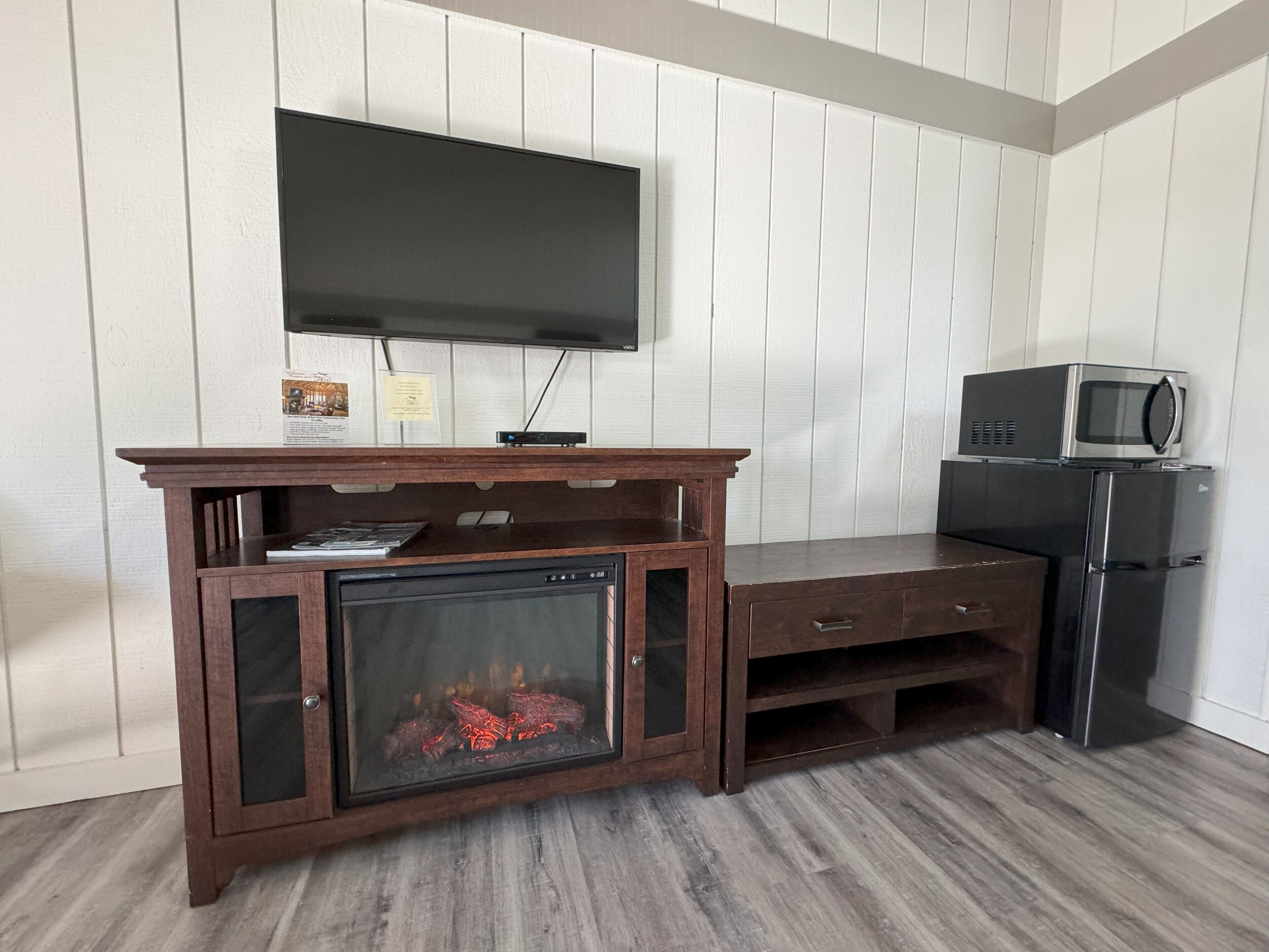 A living room with a wooden entertainment center, a flat-screen TV mounted above it, a built-in fireplace, a microwave, and a refrigerator.