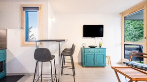 A modern kitchen with a dining area, a TV mounted on the wall, and a view of greenery outside.