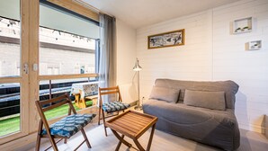 A modern living room with a sofa, two wooden chairs, a small table, a floor lamp, and a view through large windows.