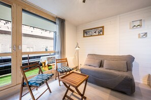 Ein modernes Wohnzimmer mit einer Couch, zwei Holzstühlen, einem kleinen Tisch, einer Stehlampe und Blick durch große Fenster.