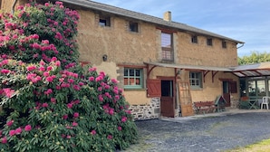 Un bâtiment rustique en pierre, doté d’un porche, d’un grand arbuste aux fleurs roses et d’une allée gravillonnée.