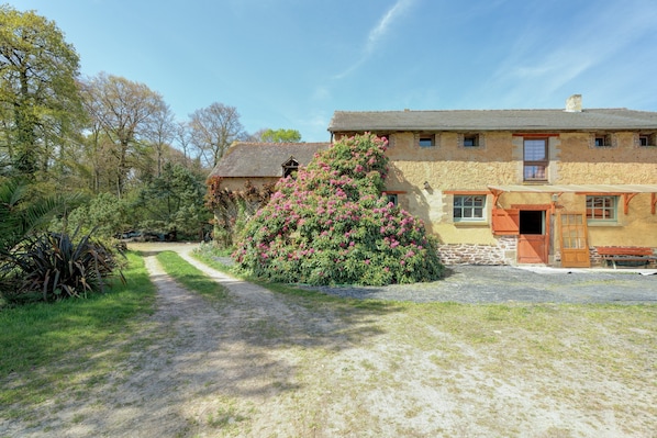A stone building with a garden path leading to a wooden door, surrounded by trees and shrubs.