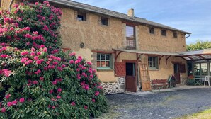 Un bâtiment rustique en pierre, doté d’un porche, d’un grand arbuste aux fleurs roses et d’une allée gravillonnée.