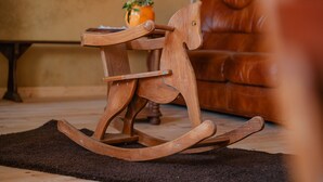Un fauteuil à bascule en bois, doté d’un coussin, est placé sur un tapis foncé dans une pièce où se trouvent un canapé en cuir marron et, à l’arrière-plan, un vase rempli de fleurs.
