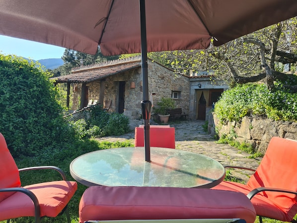 A stone building with a tiled roof, a glass table with red chairs, and a large umbrella.