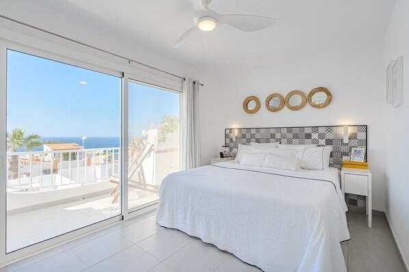A bedroom with a large bed, a ceiling fan, a nightstand, and a view of the sea.
