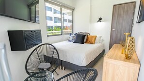 Une chambre moderne avec un lit, deux chaises noires, une petite table, un meuble en bois et un téléviseur fixé au mur.