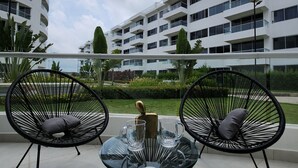 Un espace extérieur moderne, aménagé avec deux chaises à l’assise en treillis métallique noir et une table en verre, offrant une vue sur un jardin paysager et un immeuble blanc à plusieurs étages.