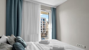 Ett modernt hotellrum med en stor säng, vita sängkläder och utsikt över byggnader genom ett fönster med blå gardiner.
