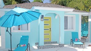 Une maison bleu clair avec une porte jaune, deux fauteuils de jardin bleus et un parasol bleu.