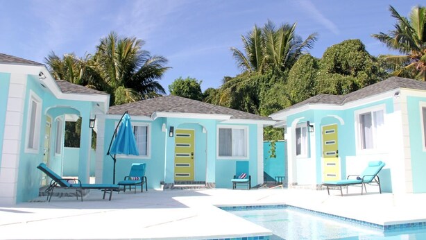 Un espace aménagé au bord de la piscine, comprenant deux petits bâtiments, chacun doté d’une porte et de fenêtres, ainsi qu’une piscine avec des chaises longues et des parasols.