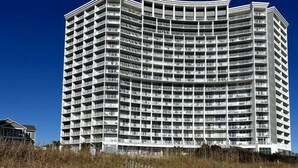 Ein mehrstöckiges Hotel mit geschwungener Fassade, direkt neben einem Sandstrand mit Holzzaun.