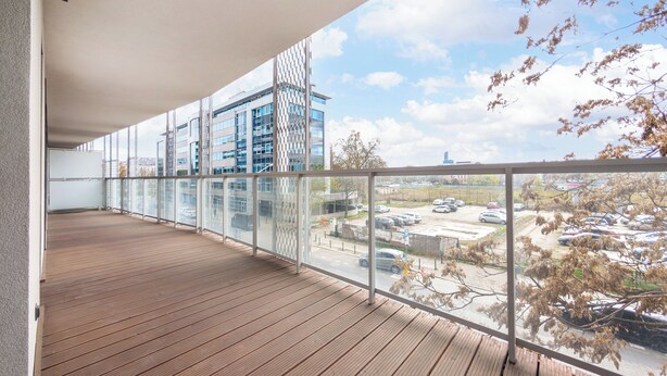 A balcony with a wooden floor and glass railings, overlooking a parking area and buildings. - Riverside Harmony Chic Apartment (Wrocław)