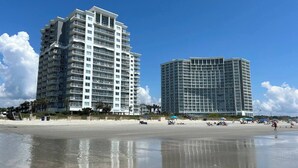Ein Strandhotel mit mehreren Etagen, ein sandiger Strand im Vordergrund und ein klarer, blauer Himmel.