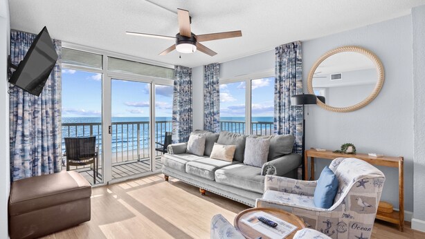 Ein geräumiges Wohnzimmer mit großem Fenster zum Meer, Deckenventilator, flachbildfernseher an der Wand und eine gemütliche Sitzecke mit einem gemusterten Sessel und Hocker.