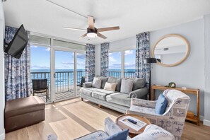 Ein geräumiges Wohnzimmer mit großem Fenster zum Meer, Deckenventilator, flachbildfernseher an der Wand und eine gemütliche Sitzecke mit einem gemusterten Sessel und Hocker.