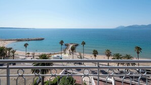 Une vue sur la côte, depuis un balcon donnant sur une plage bordée de palmiers, un brise-lames rocheux et un ciel bleu dégagé.