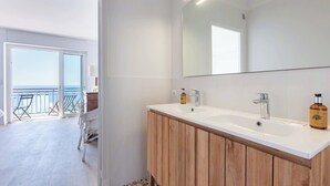 Une salle de bain moderne avec deux vasques, un grand miroir et une vue sur la mer, visible à travers une porte ouverte.