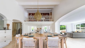 A spacious dining area with a high ceiling, a large chandelier, and a view of the outdoors through large windows.