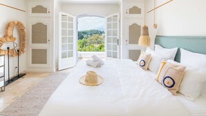 A spacious bedroom with an open door leading to a balcony, a bed with white linens, and a view of greenery.