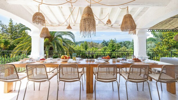 A dining area with a long table set for six, surrounded by wicker chairs, under a white ceiling with hanging lights.