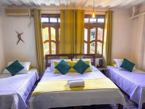 Habitación de hotel con dos camas, cada una con cubrecama blanco acolchado y almohadas color turquesa. Se ve una ventana con cortinas amarillas, una lámpara colgante y un aire acondicionado de pared.