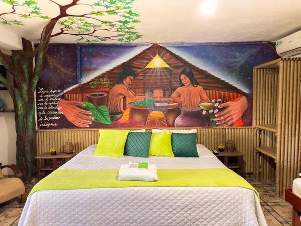 A room with a mural depicting indigenous people, a bed with green and white bedding, and a tree-like structure.