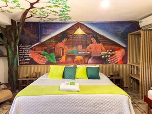 Una habitación con un mural que representa a indígenas, una cama con ropa de cama verde y blanca, y una estructura similar a un árbol.