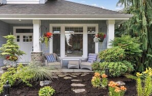 Enjoy a relaxing moment on the porch with a seating area, surrounded by lush greenery and a stone pathway leading to the entrance.