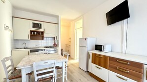 A compact kitchen with white cabinets, a wooden dining table, and a flat-screen TV mounted on the wall.