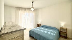 A bedroom with a bed, a wooden dresser, a ceiling fan, and a window with sheer curtains.
