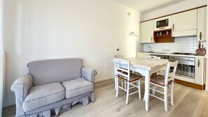 A compact kitchen with white cabinets, a wooden countertop, and a dining area with a checkered sofa and white chairs.