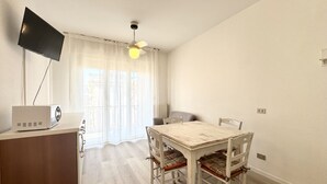 A small, well-lit room with a wooden dining table, white chairs, a ceiling fan, a flat-screen TV mounted on the wall, and a window with sheer curtains.